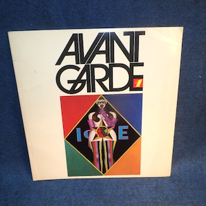 May include: A white book cover with the title "AVANT GARDE" in black bold letters. The cover features a colorful illustration of a woman in a purple glove and sunglasses standing in a diamond-shaped frame.