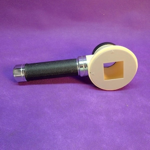 May include: A medical dermatoscope with a black textured handle, silver accents, and a cream-colored head with a square opening. The device is designed for skin examination and diagnosis.