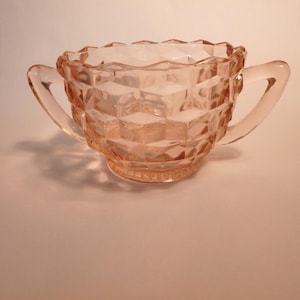 May include: A vintage pink glass sugar bowl with two handles. The bowl has a geometric, diamond-like pattern and a scalloped rim. The sugar bowl is made of clear glass and has a light pink hue.