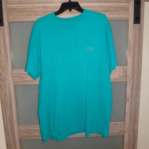 May include: A turquoise short-sleeved t-shirt with the IBM logo on the chest. The shirt is hanging on a black hanger against a wooden door background. The shirt is a solid color and appears to be made of cotton.