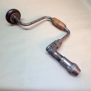 May include: A vintage hand drill with a wooden handle and a dark brown knob. The metal drill has a curved design and a chuck for holding drill bits. The tool shows signs of wear and age.