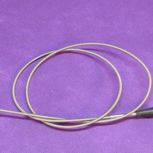 May include: A flexible silver-toned metal cable release for a camera, featuring a coiled wire body, a threaded metal tip at one end, and a black plastic plunger mechanism with a small side screw at the other end for shutter activation.