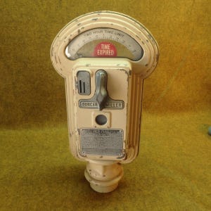 May include: A vintage beige Duncan Miller parking meter with a curved top display showing the text TWO HOUR TIME LIMIT and a red indicator reading TIME EXPIRED. The metal housing features a coin slot, a central rotating handle, and a metal plate.