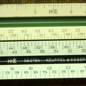 May include: A Keuffel & Esser slide rule featuring a black central bar with the text K&E 563784 KEUFFEL & ESSER, flanked by off-white scales with black numerical markings and a green grooved section, showing imperial inch measurements.