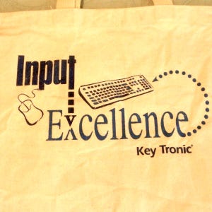 May include: Cream-colored tote bag with the words "Input Excellence" in dark blue, along with a mouse and keyboard graphic. The words "Key Tronic" are printed below the main text.
