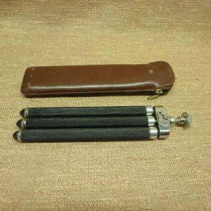 May include: A vintage black and silver tripod with a brown leather case. The tripod has three legs with a textured grip and silver accents. The case is rectangular with a zipper closure.