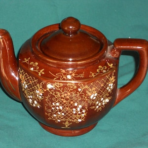 May include: A brown ceramic teapot with a lid and a floral design in gold and white. The teapot has a rounded body and a spout on the side.