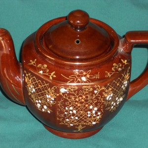 May include: A brown ceramic teapot with a lid and a gold and white floral design.