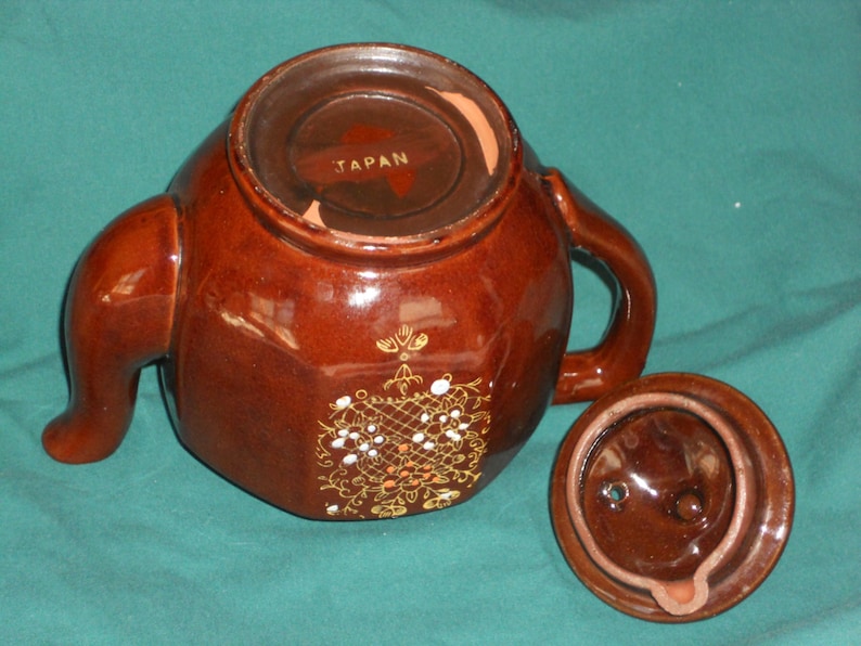 May include: A brown ceramic teapot with a gold floral design and a lid. The teapot is marked "JAPAN" on the bottom.