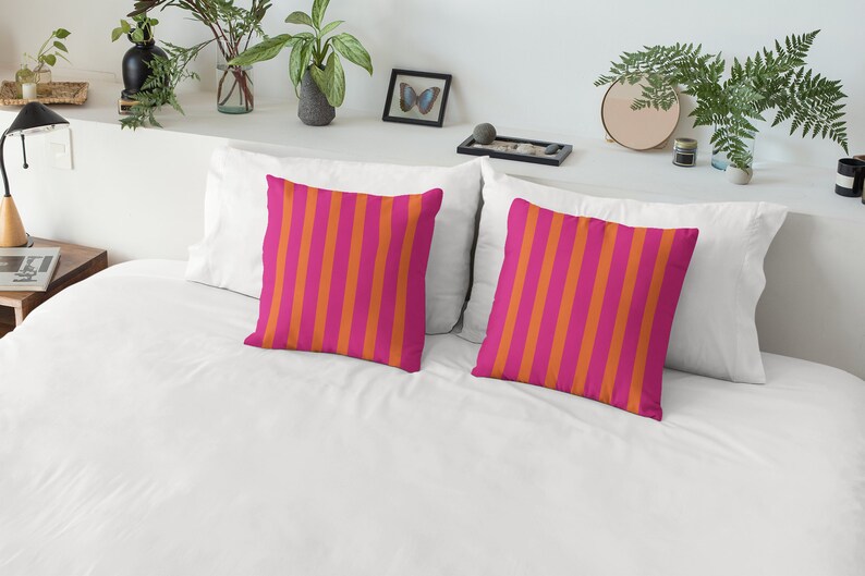 May include: Two decorative throw pillows with a pink and orange striped pattern. The pillows are on a white bed with a white headboard.