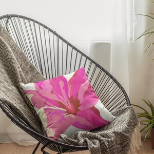 Op de afbeelding: Een zwarte metalen stoel met een roze bloemenkussen. De stoel staat in een kamer met een witte muur en een raam met witte gordijnen. Een groene plant staat in een bruine pot op de vloer.