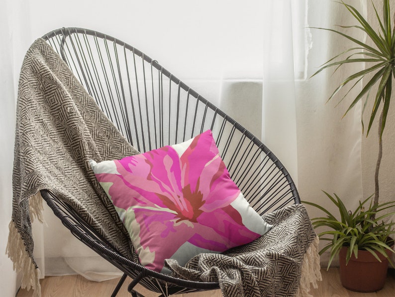 Op de afbeelding: Een zwarte metalen stoel met een roze bloemenkussen. De stoel staat in een kamer met een witte muur en een raam met witte gordijnen. Een groene plant staat in een bruine pot op de vloer.