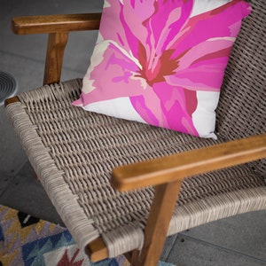 Op de afbeelding: Een roze bloemenkussen met een witte achtergrond ligt op een gevlochten rieten stoel met houten armleuningen. De stoel staat op een tapijt met een patroon.