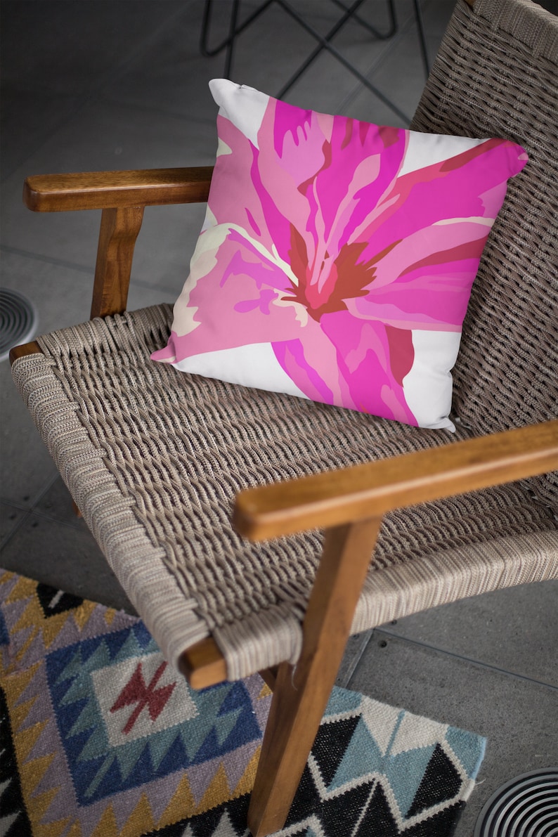 Op de afbeelding: Een roze bloemenkussen met een witte achtergrond ligt op een gevlochten rieten stoel met houten armleuningen. De stoel staat op een tapijt met een patroon.