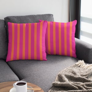 May include: Two pink and orange striped throw pillows on a grey sofa with a black and white patterned throw blanket and a wooden side table with a white coffee cup and saucer.