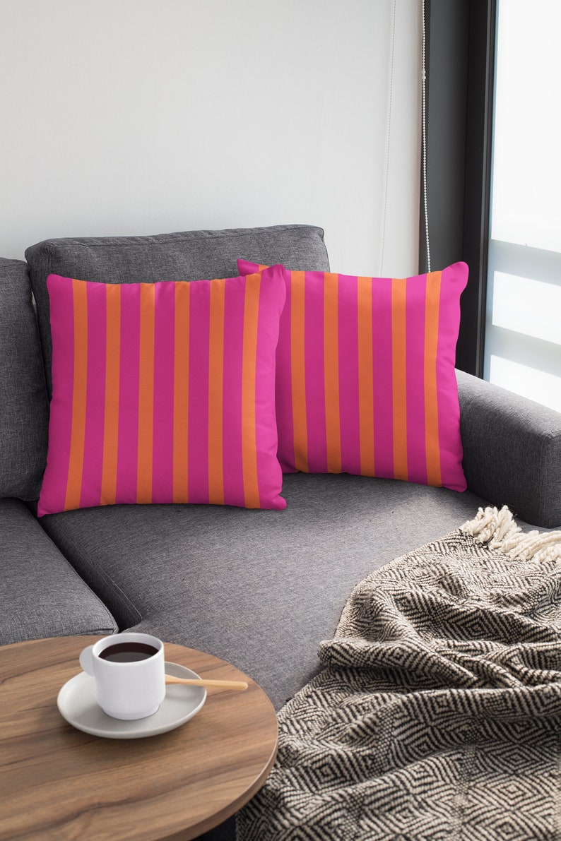May include: Two pink and orange striped throw pillows on a grey sofa with a black and white patterned throw blanket and a wooden side table with a white coffee cup and saucer.