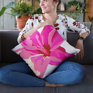 Op de afbeelding: Een vrouw die op een grijze bank zit en een roze bloemenkussen vasthoudt. Het kussen heeft een witte achtergrond met een grote roze bloem in het midden.