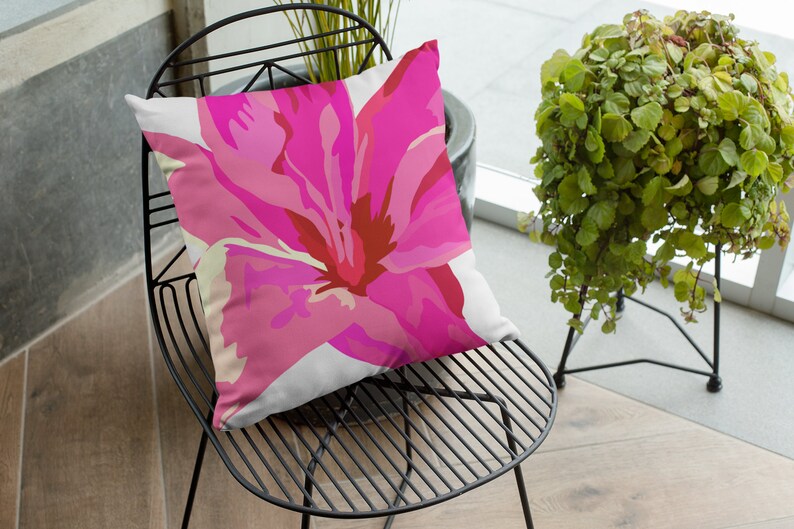 Op de afbeelding: Een roze bloemen kussen met een witte achtergrond. Het kussen ligt op een zwarte metalen stoel.