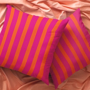 May include: Two decorative throw pillows with a pink and orange striped pattern. The pillows are on a peach-coloured fabric surface.