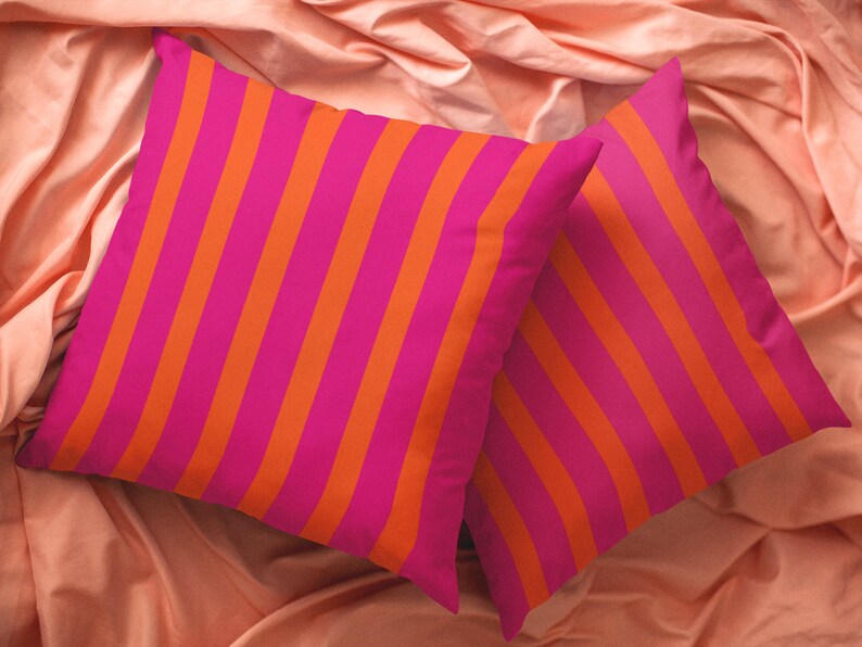 May include: Two decorative throw pillows with a pink and orange striped pattern. The pillows are on a peach-coloured fabric surface.