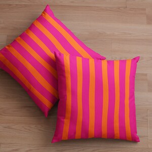 May include: Two decorative throw pillows with a pink and orange striped pattern. The pillows are lying on a wooden floor.