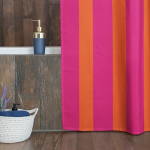 May include: A pink and orange striped shower curtain hangs in a bathroom with a white bath and a wooden wall. A white wicker basket with a blue towel and lavender flowers sits on the floor.
