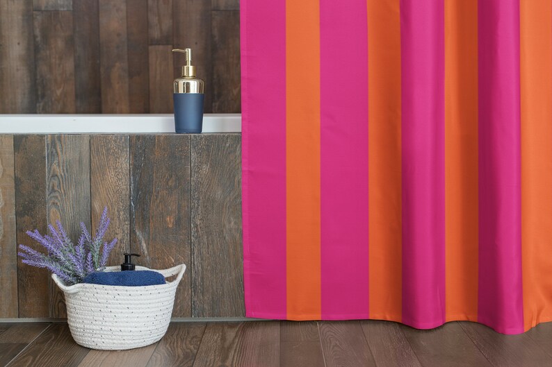 May include: A pink and orange striped shower curtain hangs in a bathroom with a white bath and a wooden wall. A white wicker basket with a blue towel and lavender flowers sits on the floor.