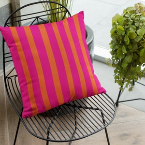May include: A pink and orange striped cushion on a black metal chair. The chair is in a room with a window and a plant.
