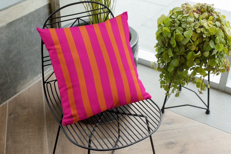 May include: A pink and orange striped cushion on a black metal chair. The chair is in a room with a window and a plant.