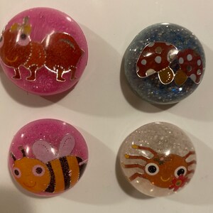 May include: Four round resin cabochons with different designs. The first cabochon is pink with a red ant with a gold crown. The second cabochon is blue with two red mushrooms with white spots. The third cabochon is pink with a yellow and black bee with pink wings. The fourth cabochon is white with a brown spider with black eyes and a red flower.