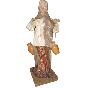 May include: A handmade paper mache figurine of a woman carrying two jugs on a stick. The woman is wearing a brown and white dress with a floral pattern. The jugs are brown and have a rustic look.