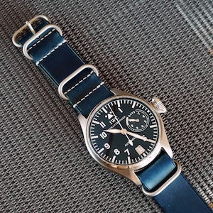 May include: A silver wristwatch with a black face and white markings. The watch has a blue leather strap with white stitching and silver buckle.