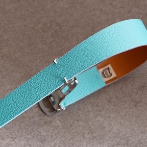 May include: A turquoise leather belt with a silver buckle. The belt has a textured surface and a contrasting orange-brown interior. The buckle is silver and the belt is curved.
