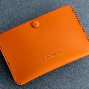 Peut inclure: Portefeuille en cuir orange avec une fermeture à bouton pression et le texte "Atelier Du Cuir" en relief sur le devant.