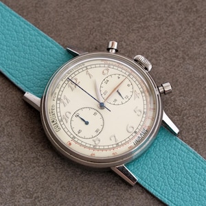 May include: A silver-toned chronograph watch with a cream-colored face and a light blue leather band. The watch face has black and blue hands, and several sub-dials. The band is approximately 2.5 cm wide.