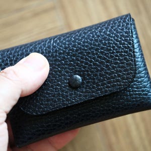 Peut inclure: Un porte-cartes en cuir noir texturé, avec une fermeture à bouton-pression. Le petit portefeuille est rectangulaire avec des bords arrondis et une surface grainée. Il est tenu dans une main.