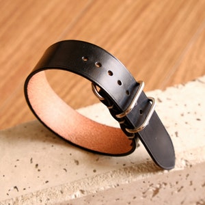 May include: A black leather watch strap with silver buckle and stitching. The strap is on a light gray textured surface.