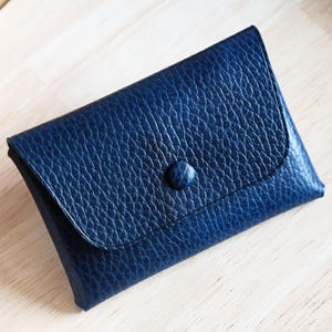 Peut inclure: Un porte-cartes en cuir bleu marine avec une surface texturée et une fermeture à bouton. Le petit portefeuille est rectangulaire et possède un rabat sécurisé par un bouton assorti. Le porte-cartes est sur une surface en bois clair.