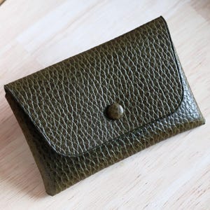 Peut inclure: Porte-cartes en cuir vert olive avec une surface texturée et une fermeture à bouton. Le petit portefeuille est rectangulaire et possède un rabat incurvé. Le porte-cartes est sur une surface en bois clair.