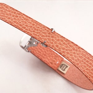May include: A salmon-colored watch band with a textured, leather-like appearance. The band features a silver-colored buckle and a small, rectangular metal piece with an embossed design. The band is angled, showcasing its details.