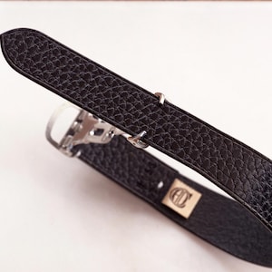 Könnte beinhalten: Schwarzes Leder-Armband mit strukturierter Oberfläche. Das Armband verfügt über eine silberfarbene Schnalle und zwei silberne Schlaufen. Eine kleine, rechteckige goldfarbene Platte mit Logo ist am Armband befestigt.