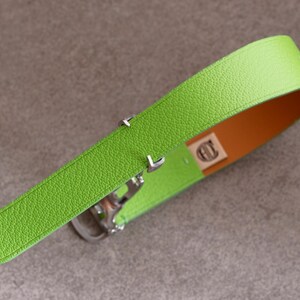 May include: A bright green leather belt with a textured surface. The interior is a contrasting orange color. The belt has a silver-colored buckle and hardware. A small tag with a logo is visible.