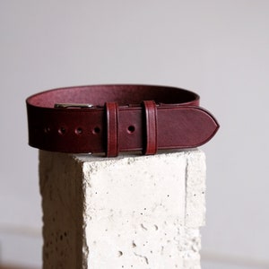 May include: A burgundy leather belt with a silver buckle. The belt is displayed on a white, textured surface. The belt has a classic design with a rounded end and several holes for adjustment.