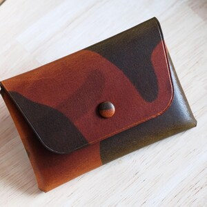 Peut inclure: Un portefeuille en cuir fait main avec un motif camouflage dans des tons de marron, orange et vert. Le portefeuille a une fermeture à rabat sécurisée par un bouton rond marron. Il est conçu pour transporter des cartes ou de l'argent.