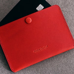 Peut inclure: Un porte-cartes en cuir rouge avec une fermeture à bouton et le texte "Atelier Du Cuir - Le Cuir Francais" en relief sur le devant.