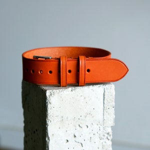 May include: A bright orange leather watch strap with a silver buckle. The strap is on a concrete block.