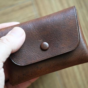 Peut inclure: Un portefeuille en cuir marron avec une fermeture à bouton-pression. Le portefeuille est rectangulaire avec des bords arrondis et un rabat. Le cuir a un aspect texturé. Mesure environ 10 cm de long.