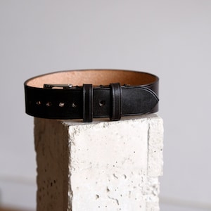 May include: A black leather belt with a silver buckle. The belt is on a white concrete block.