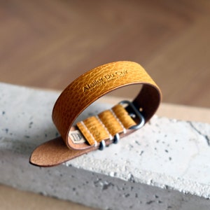 May include: A yellow leather watch strap with a silver buckle. The strap is embossed with the text "Atelier Du Cuir Handmade in France".