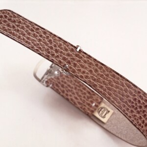 May include: A brown leather watch strap with a silver buckle. The strap has a textured surface.